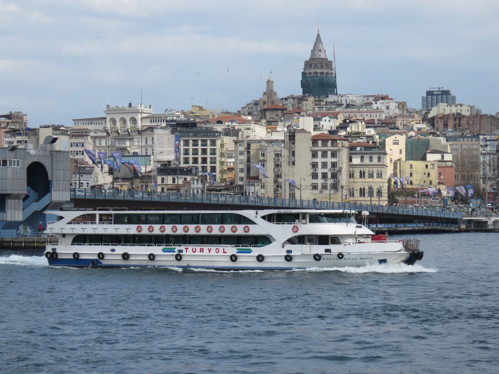 Galata Bridge seen from the water with a ferry passing and the Galata Tower on the hill behind