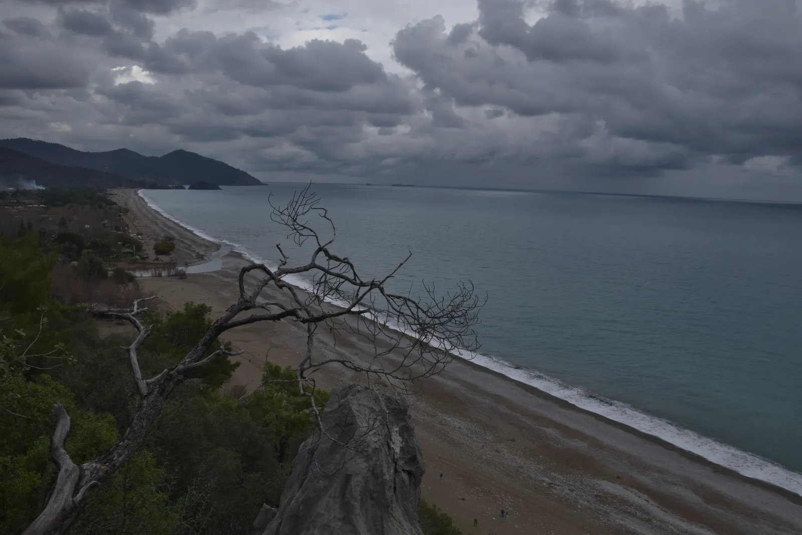 Olympos Beach seen from the headland with pebble shore, clear water, and the forested river valley behind